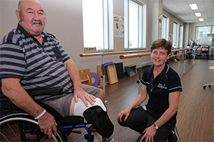 An older man whose right leg has been amputated below the knee sits in a wheelchair in a gymnasium. A female physiotherapist kneels beside the wheelchair.