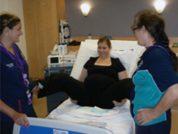 A fully clothed pregnant woman sits on a bed with her legs wide apart in a supported sitting position for labour. A female health processional stands beside her feet on each side of the bed.