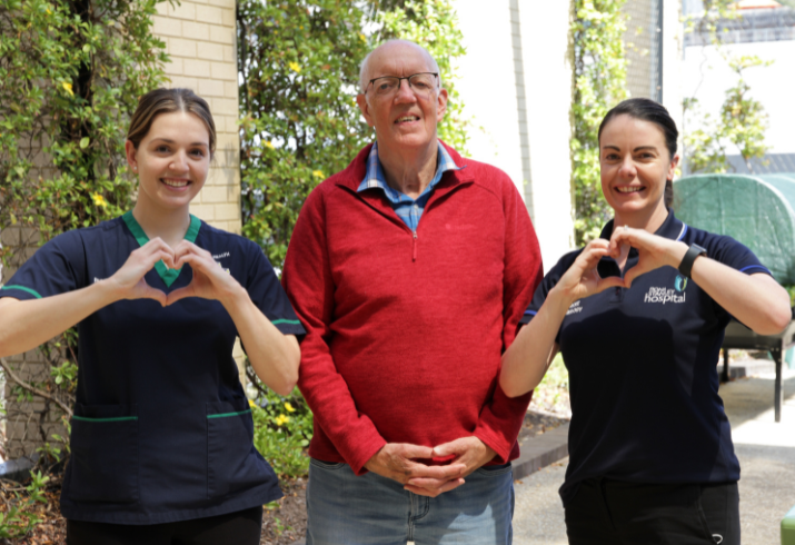 An older man stands between two female allied health professionals. The two women are making a heart gesture with their hands.