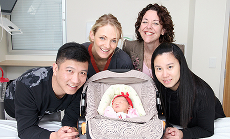 A young man and woman are beside a newborn baby in a capsule that is resting on a hospital bed. A female nurse and another woman stand with them.