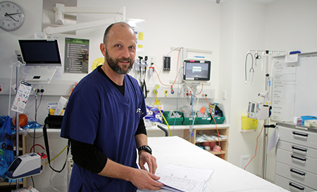 A man wearing theatre scrubs stands in a room with medical equipment.