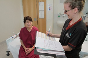 A woman in pyjamas sitting on a bed while a nurse holding a clipboard stands beside her
