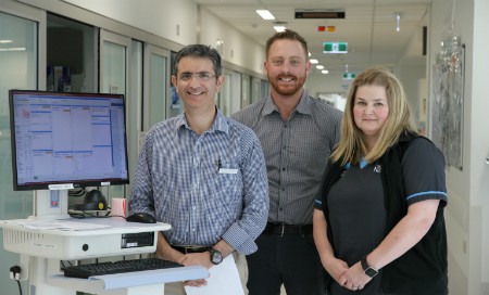 Three staff members standing next to computer on wheels in a ward setting