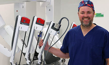 A man wearing theatre scrubs stands beside a piece of medical equipment.