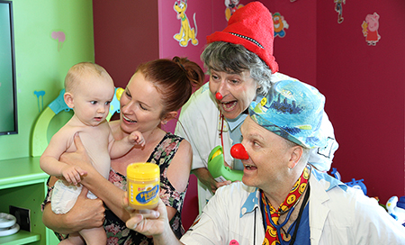 A woman holds a young baby. A man and woman dressed as clown doctors are standing next to them