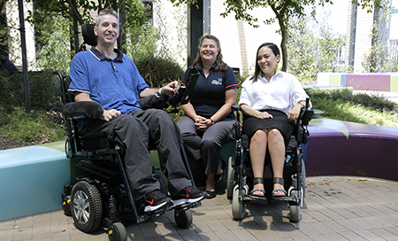 A man and woman, both seated in wheelchairs, in an outdoor courtyard. Another women sits with them on a brightly coloured seat.
