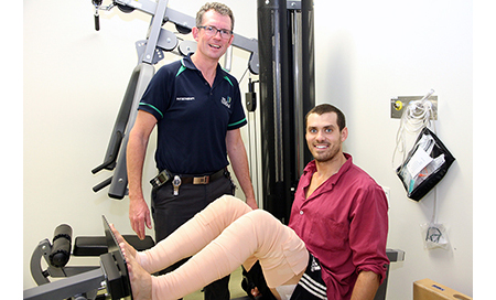 A male physiotherapist stands beside a young man on a piece of fitness equipment 