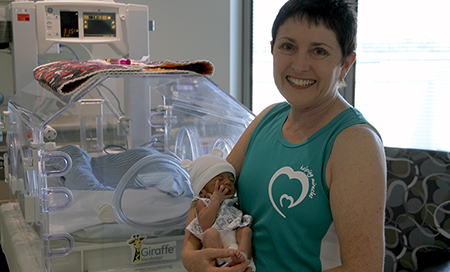 A woman standing in front of an incubator holds a newborn baby. 