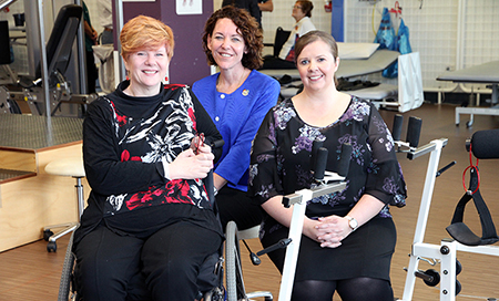 Three women are seated in a gym therapy area. One of the women is seated in a wheelchair.